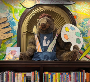 photo of a teddy bear dressed as an artist sitting on top of a library bookcase